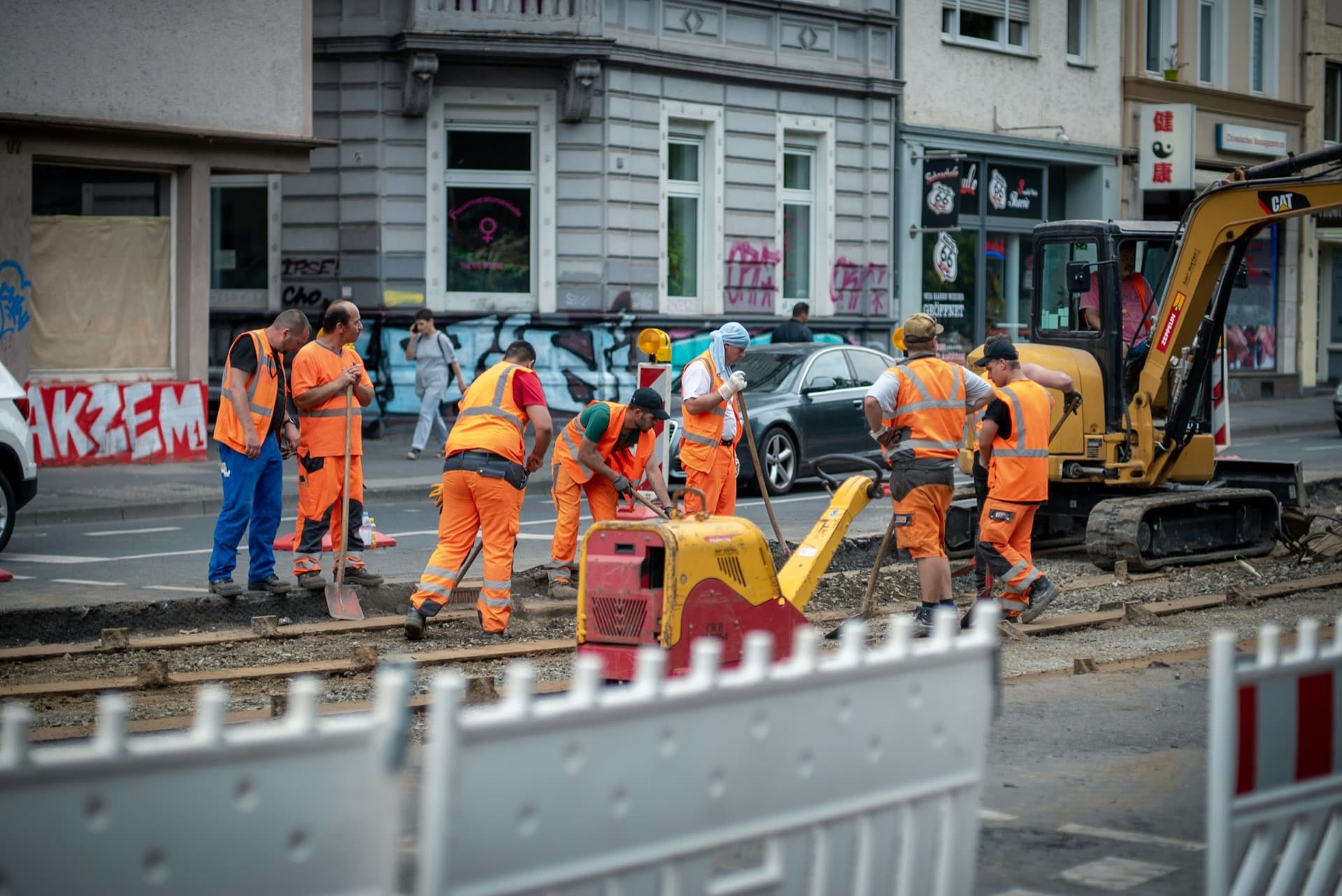Sept ouvriers en tenue orange sur un chantier de voirie en ville, autour d’une plaque vibrante et d’une mini-pelle